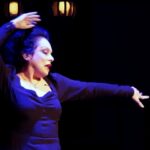 Singer Eleanora Owen wears a black dress on stage with her left had extended to the side and her right hand lifted above her head on a black stage with two back lights illuminating her