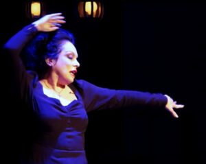 Singer Eleanora Owen wears a black dress on stage with her left had extended to the side and her right hand lifted above her head on a black stage with two back lights illuminating her