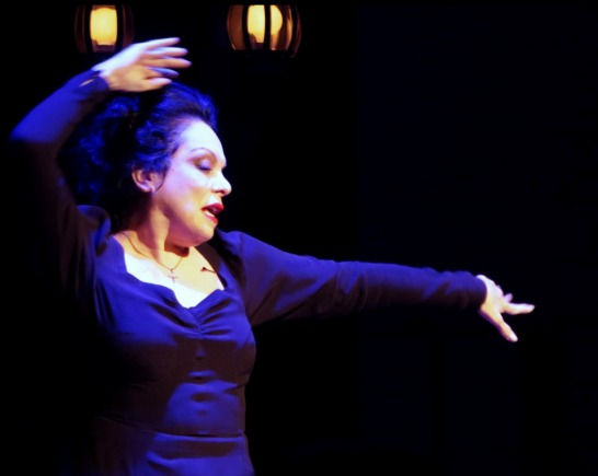 Singer Eleanora Owen wears a black dress on stage with her left had extended to the side and her right hand lifted above her head on a black stage with two back lights illuminating her