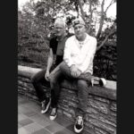 Black and white photo of Two men sitting together on a rock wall with trees in background