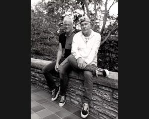 Black and white photo of Two men sitting together on a rock wall with trees in background