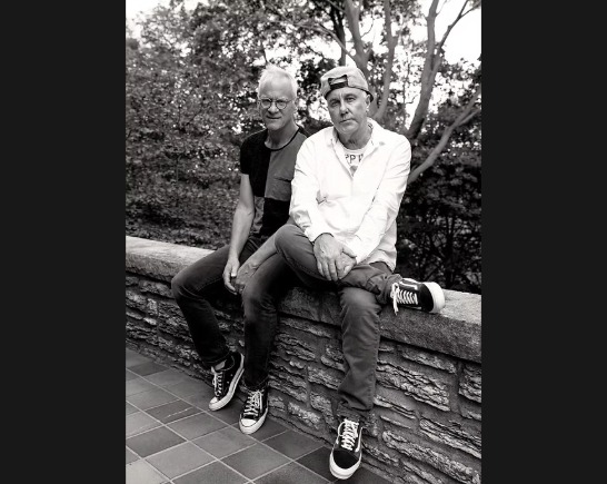 Black and white photo of Two men sitting together on a rock wall with trees in background