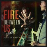 Music single cover for Sons of Silver song Fire Between Us in reds and deep browns. The keyboard player faces the left and the guitar player holds out his hand at the center of the image as if to stop us