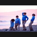Four non-binary people wear khaki pants and blue oxford shirts as they stand on a beach with a beautiful pink sky behind them