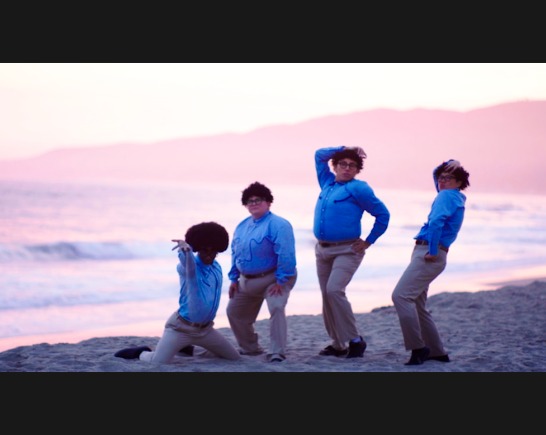 Four non-binary people wear khaki pants and blue oxford shirts as they stand on a beach with a beautiful pink sky behind them