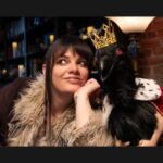 Cassie Workman in her studio wears a coat with a fur collar she smiles looking up to the right as a crow with a crown and red eyes leans against her on her left