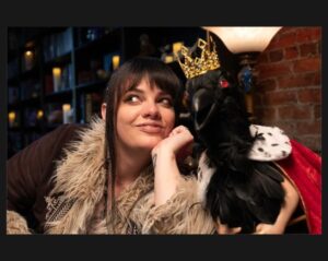 Cassie Workman in her studio wears a coat with a fur collar she smiles looking up to the right as a crow with a crown and red eyes leans against her on her left