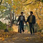 a man and woman dressed in pacific northwest style walk hand in hand along path bordered by trees with golden autumn leaves
