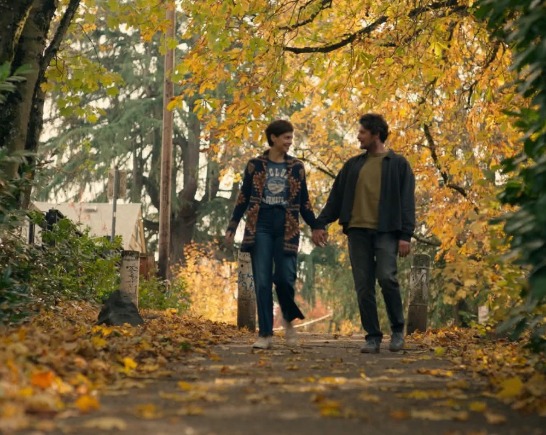 a man and woman dressed in pacific northwest style walk hand in hand along path bordered by trees with golden autumn leaves