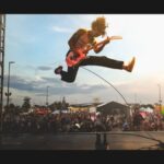 A thin rock n roll musician is onstage at an outdoor festival with the crowd in the background. He is airborne with both legs outstretched as he plays a red electric guitar