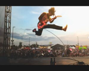 A thin rock n roll musician is onstage at an outdoor festival with the crowd in the background. He is airborne with both legs outstretched as he plays a red electric guitar