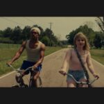 two adolescent siblings ride bicycles on a country road. The girl is white with blonde hair. She wears a pink sleeveless shirt and jean shorts The boy is black and wears a grey stocking cap, jean shorts and a white tank top