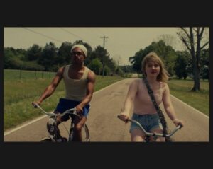 two adolescent siblings ride bicycles on a country road. The girl is white with blonde hair. She wears a pink sleeveless shirt and jean shorts The boy is black and wears a grey stocking cap, jean shorts and a white tank top